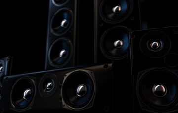 Multiple large sleek black speakers with chrome centers enhanced by accent lighting in a dark room.