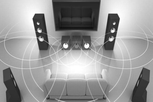 A large, sleek black flat screen TV is wall mounted in a living room along with multi large, black home theater speakers. Sound Waves are circling the room to emphasize how the sound will engulf the viewer sitting on the white couch in a movie theater-like surround sound experience!